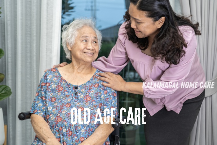 A warm interaction between an elderly woman and a caregiver from Kalaimagal Home Care in Coimbatore. The caregiver, wearing a pink blouse, gently places a comforting hand on the elderly woman's shoulder, exemplifying compassionate old age care. The elderly woman, seated and wearing a floral dress, looks up with a smile, reflecting the supportive and caring environment provided.